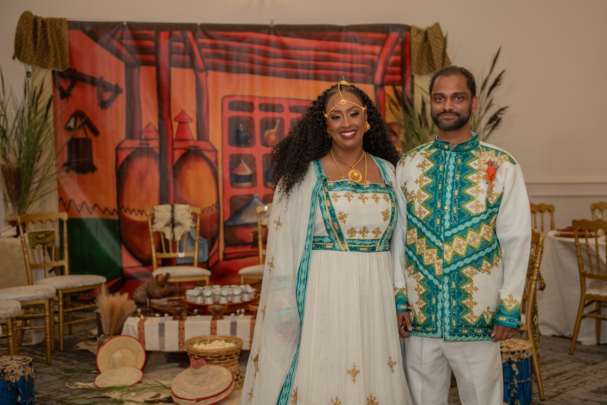 Salem and Shannon stand smiling in traditional African-inspired attire, adorned with gold jewelry and teal embroidery.