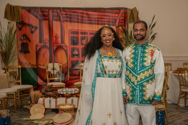 Salem and Shannon stand smiling in traditional African-inspired attire, adorned with gold jewelry and teal embroidery.