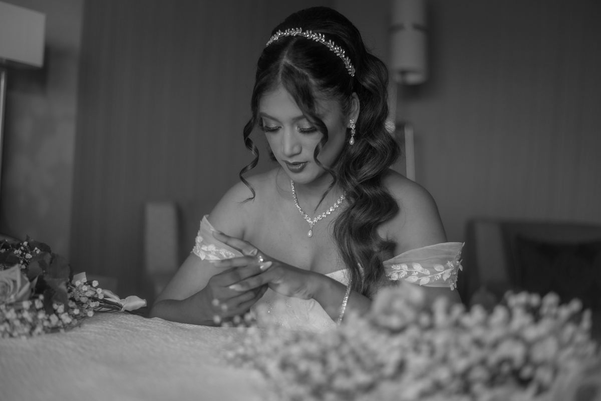 A bride in monochrome sits at a table with flowers, adjusting her wedding ring, illuminated by soft light.