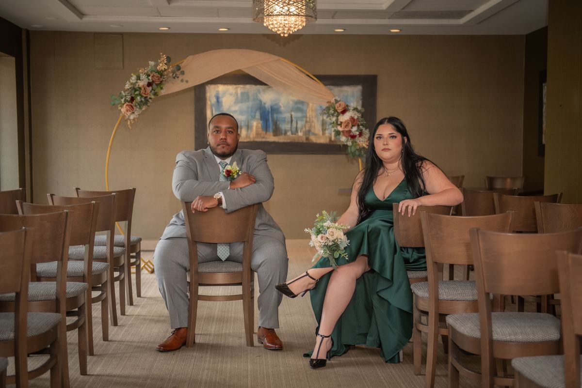 A couple in wedding attire sit in an empty ceremony space with a floral arch, looking at the camera.