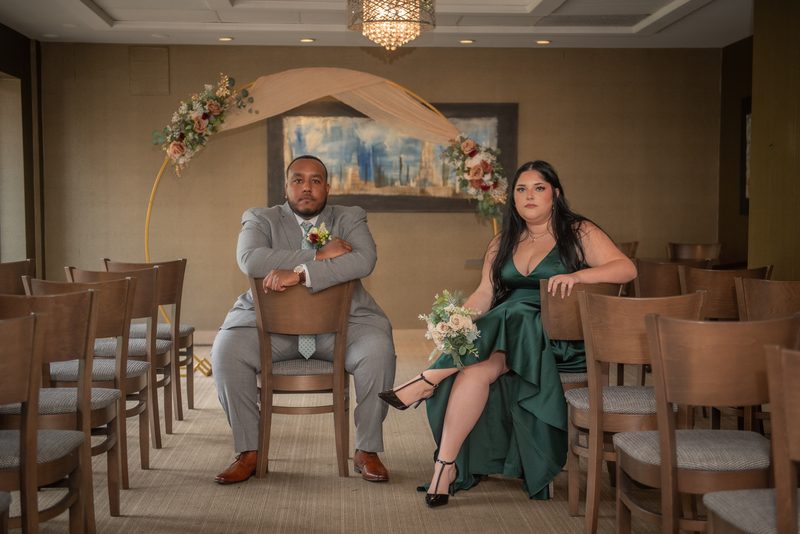 A couple in wedding attire sit in an empty ceremony space with a floral arch, looking at the camera.