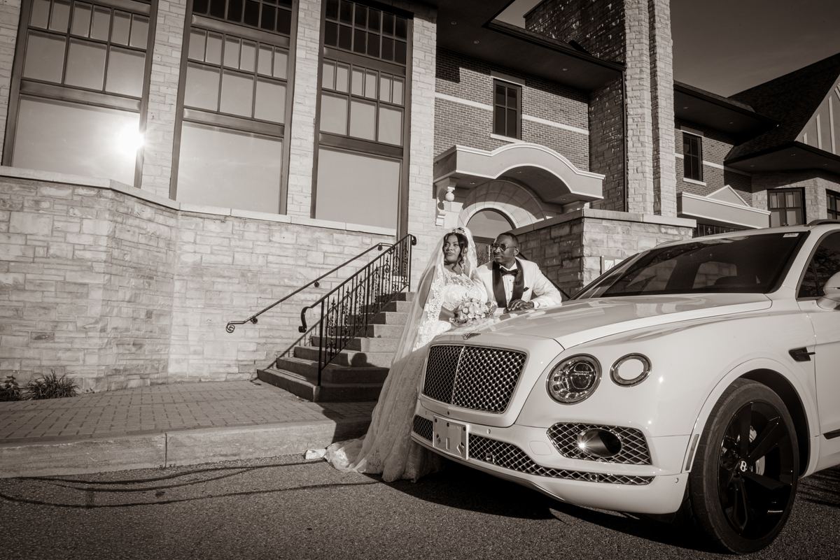 A bride and groom pose by a white car in front of an arched entryway, with dramatic black-and-white lighting and lens flare.