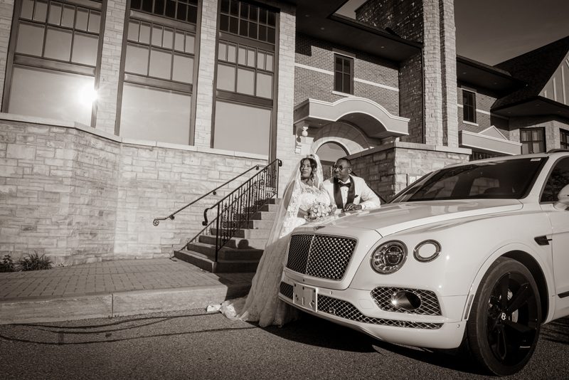 A bride and groom pose by a white car in front of an arched entryway, with dramatic black-and-white lighting and lens flare.