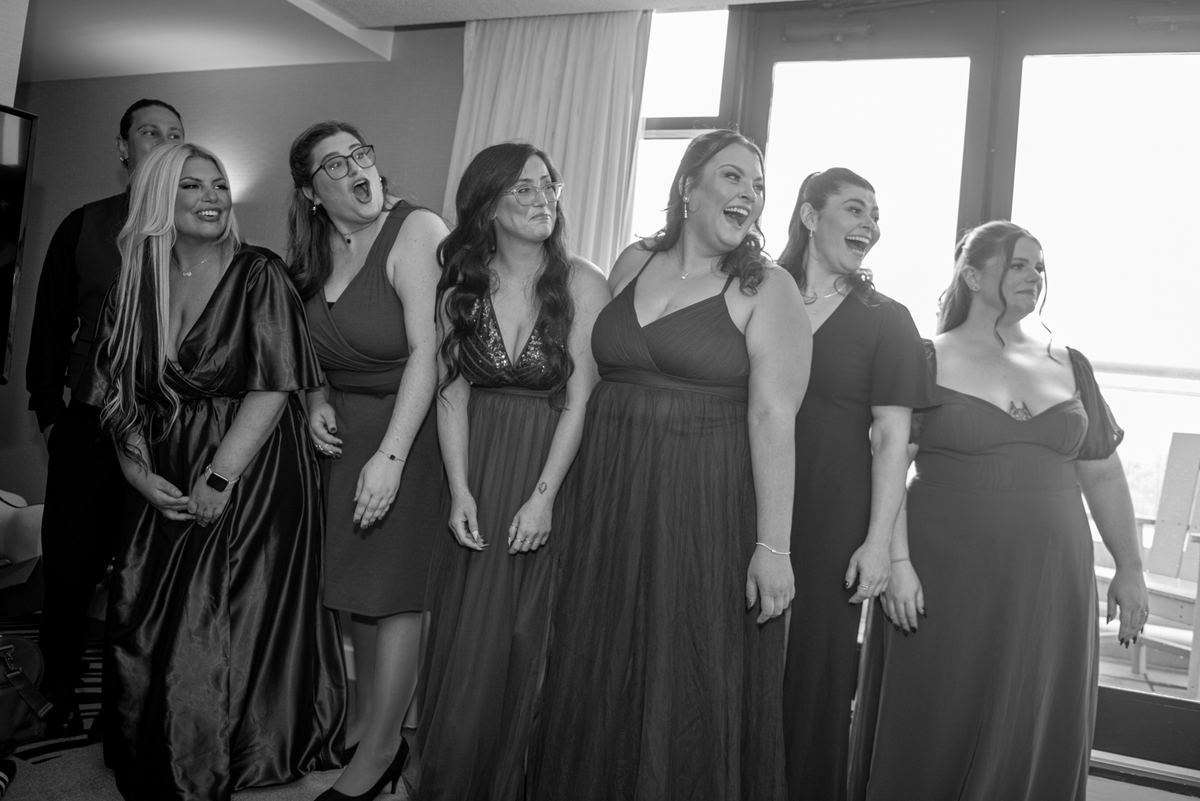 Bridesmaids in elegant evening gowns laugh together, illuminated by natural light streaming through a window.