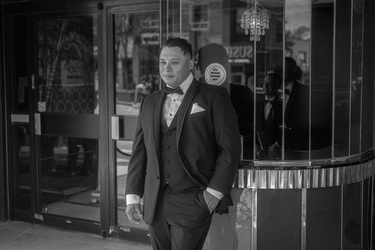 A man in a tuxedo stands outside a glass-fronted building, his reflection visible, under soft ambient light.