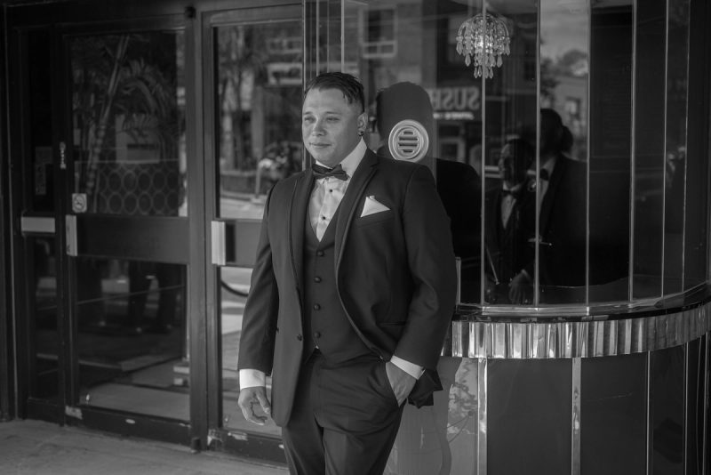 A man in a tuxedo stands outside a glass-fronted building, his reflection visible, under soft ambient light.