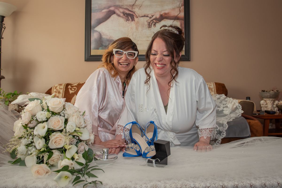 Two bridesmaids in white and pink laugh beside a table with a bouquet, blue shoes, and an open ring box.