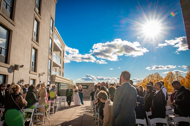 A bride and groom walk down the aisle toward an altar under a sunburst, with guests watching formally dressed on either side.