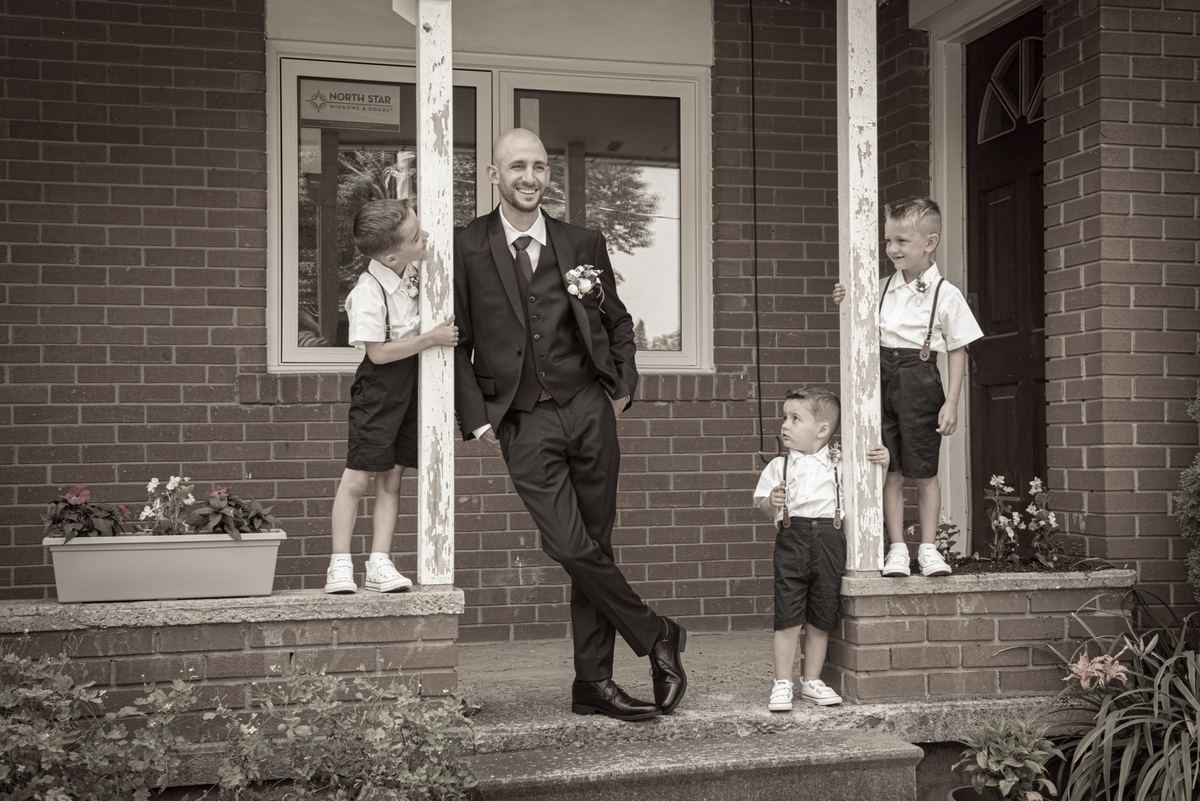 A groom in formal attire stands with three young boys dressed as page boys on a brick porch, bathed in soft daylight.
