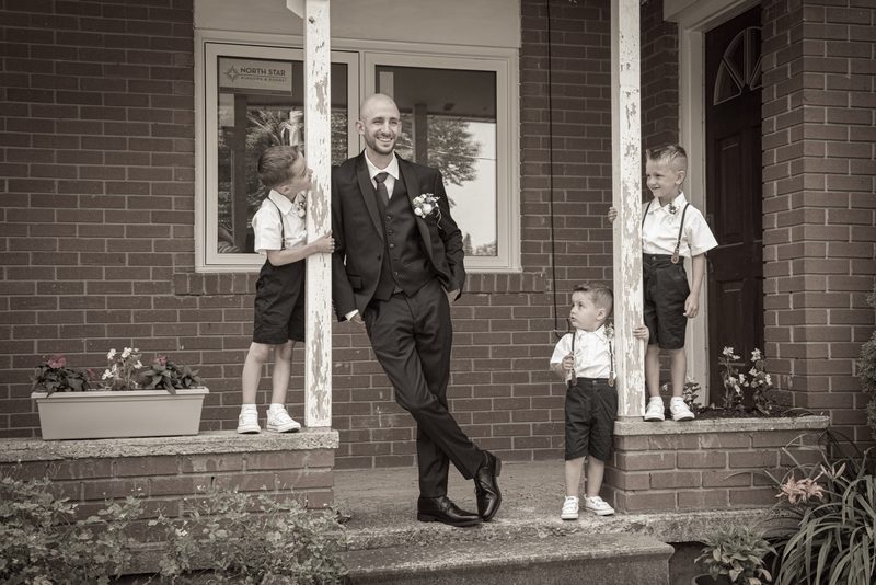 A groom in formal attire stands with three young boys dressed as page boys on a brick porch, bathed in soft daylight.