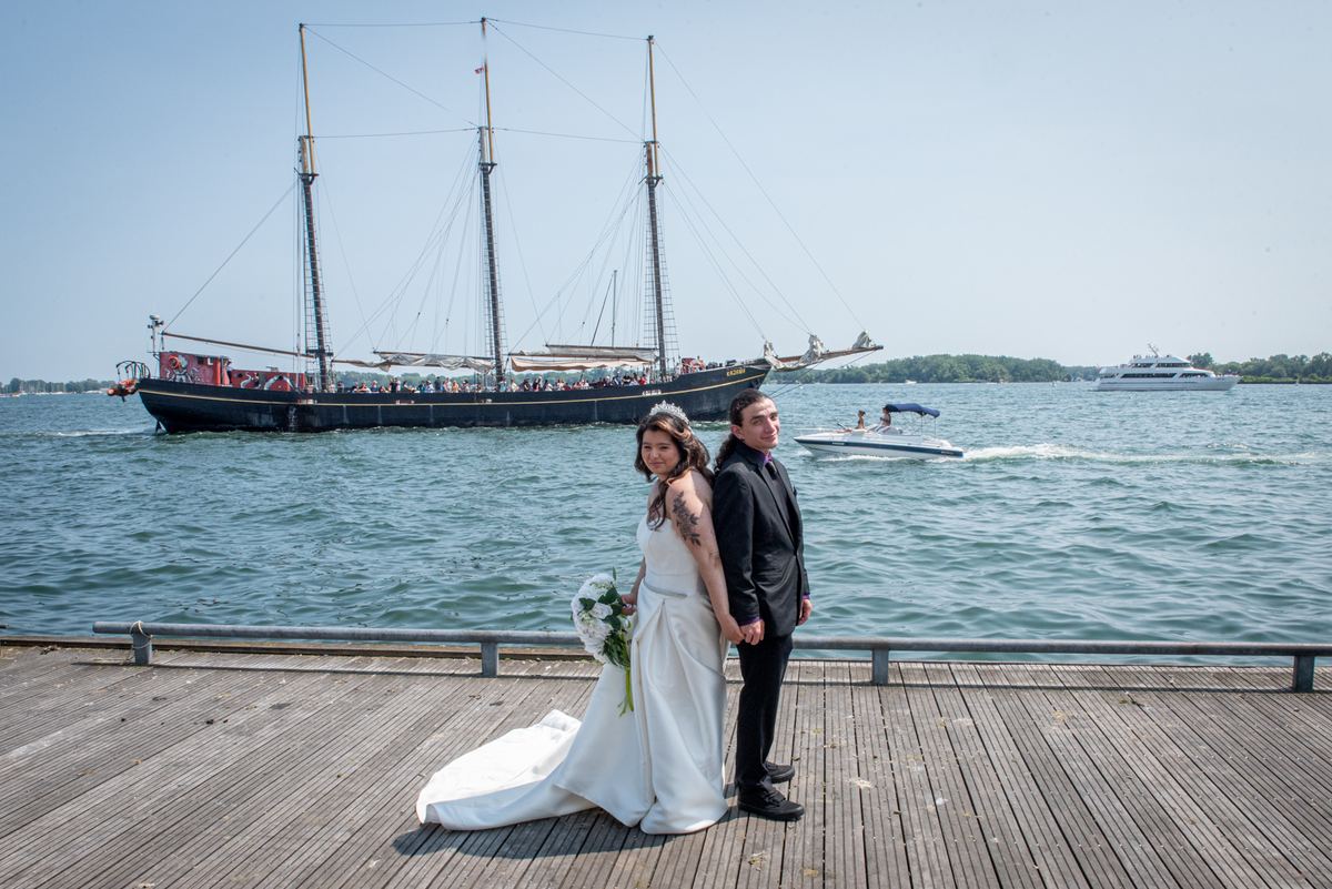 A smiling bride and groom stand on a wooden dock with sailing ships in the background during their waterfront wedding.
