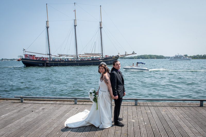 A smiling bride and groom stand on a wooden dock with sailing ships in the background during their waterfront wedding.