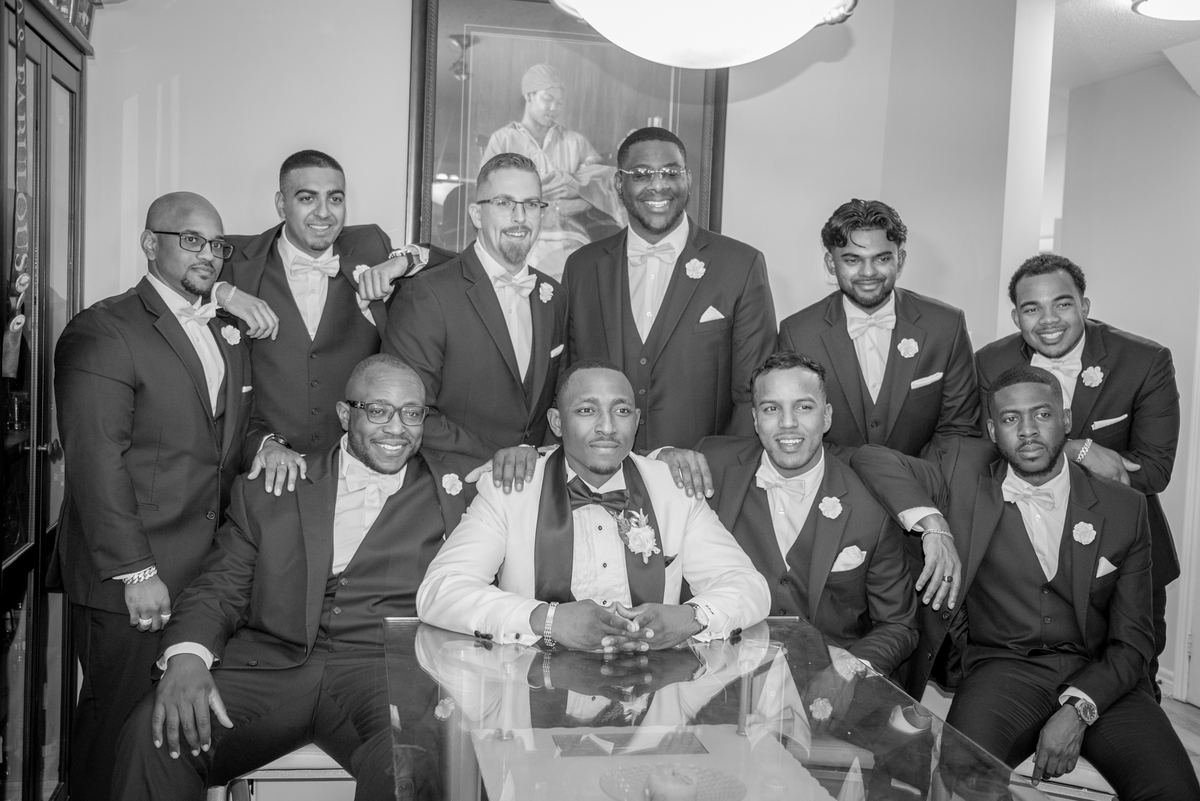 A groom and his groomsmen, dressed in matching suits with boutonnieres, smile and embrace around a glass table.
