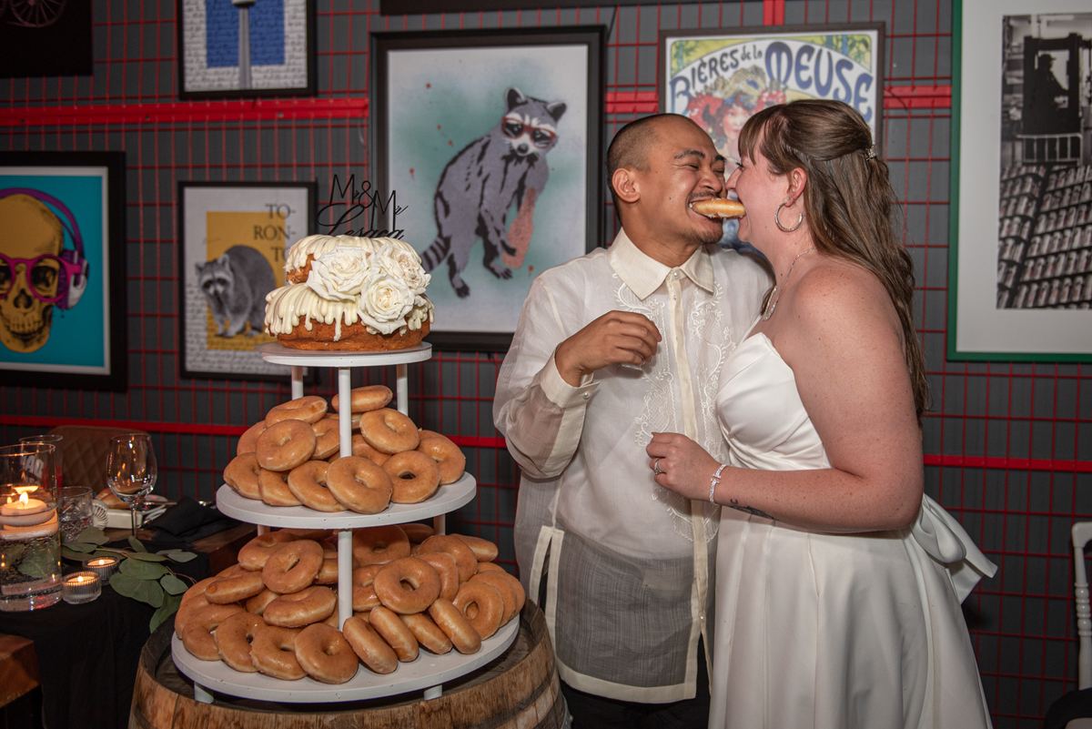 A newlywed couple laughs as they share a playful bite of their wedding cake topped with M&M and Bride & Groom toppers.