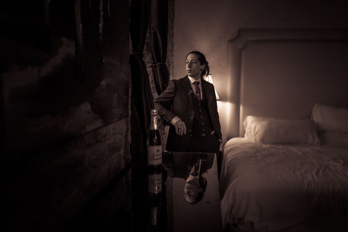 A formally dressed person stands thoughtfully in a dim hotel room, illuminated by a bedside lamp near champagne.