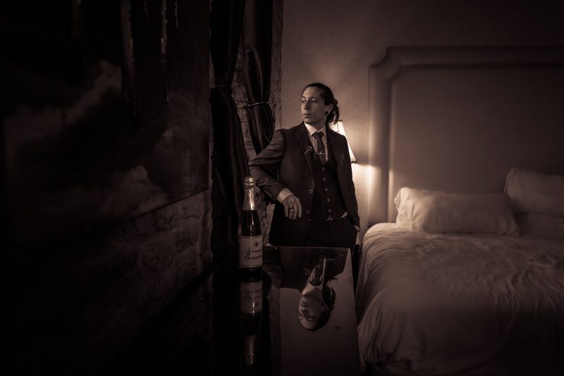 A formally dressed person stands thoughtfully in a dim hotel room, illuminated by a bedside lamp near champagne.