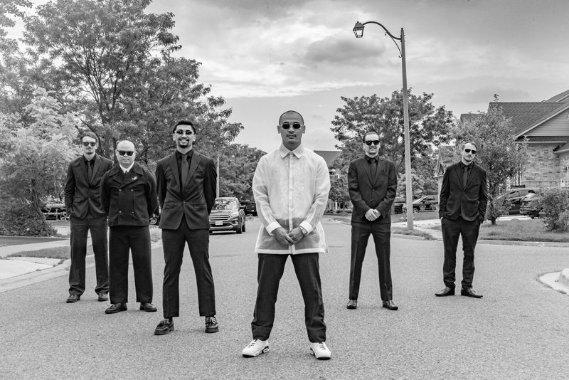 Six formally dressed men stand in a suburban street, with one wearing a light-colored barong tagalog and sneakers.