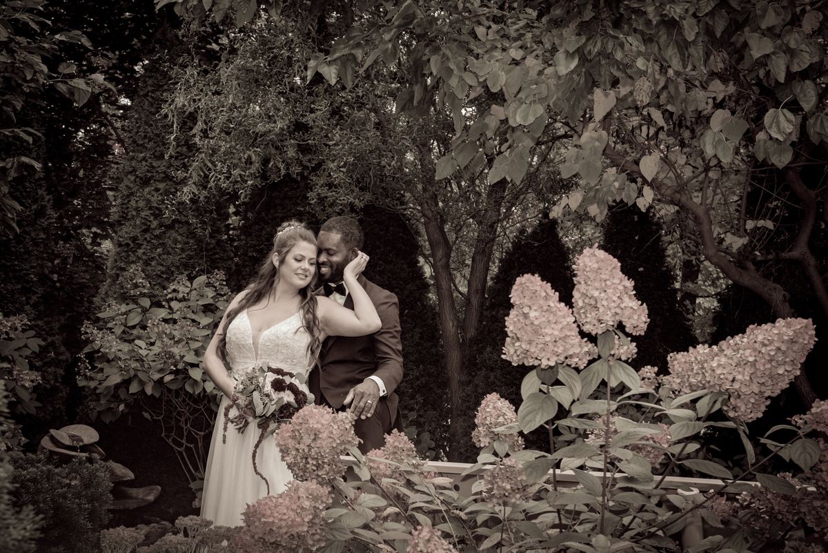 A newlywed couple embraces in a garden, the bride resting her head on the groom's shoulder amid pink hydrangeas.