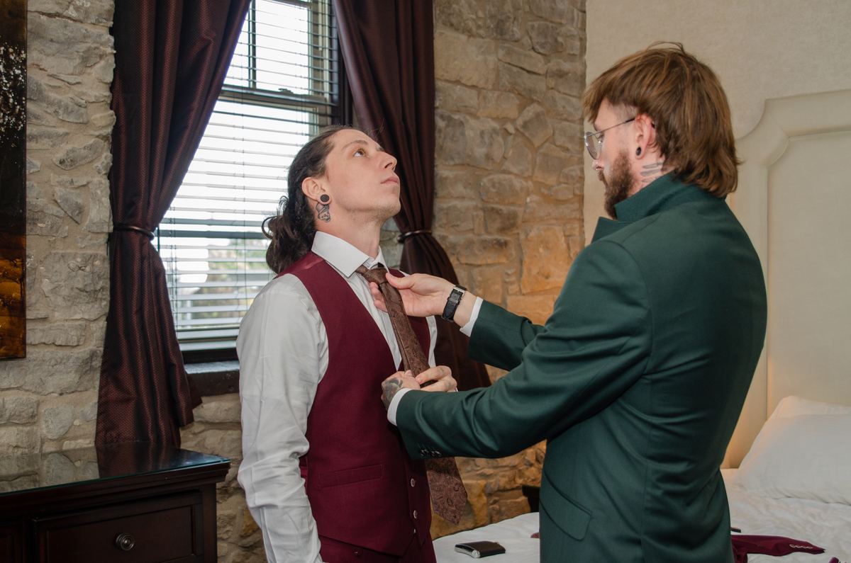 A man adjusts another's tie in a warmly lit room with stone walls and draped windows, suggesting preparation and care.