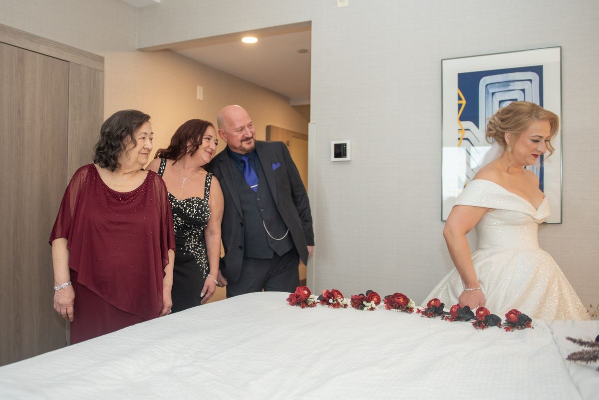 A bride in an off-the-shoulder gown stands by a bed with red and black flowers as guests smile nearby.