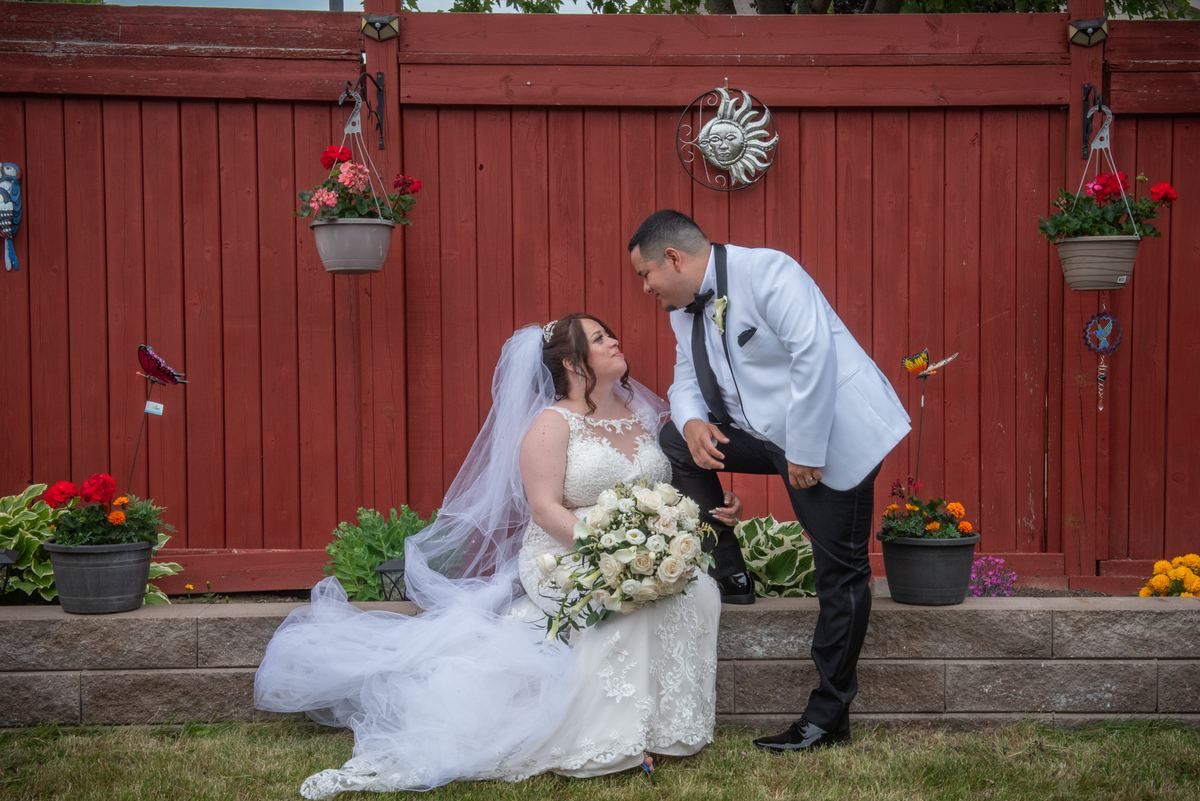Bride and groom exchange loving glances by a red fence covered in flowers during their outdoor celebration.