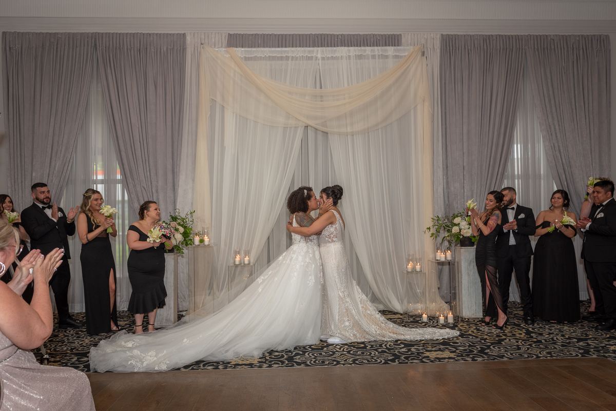 Two brides, Alexa and Nazareth, share an intimate kiss at the center of a draped archway during their indoor ceremony.