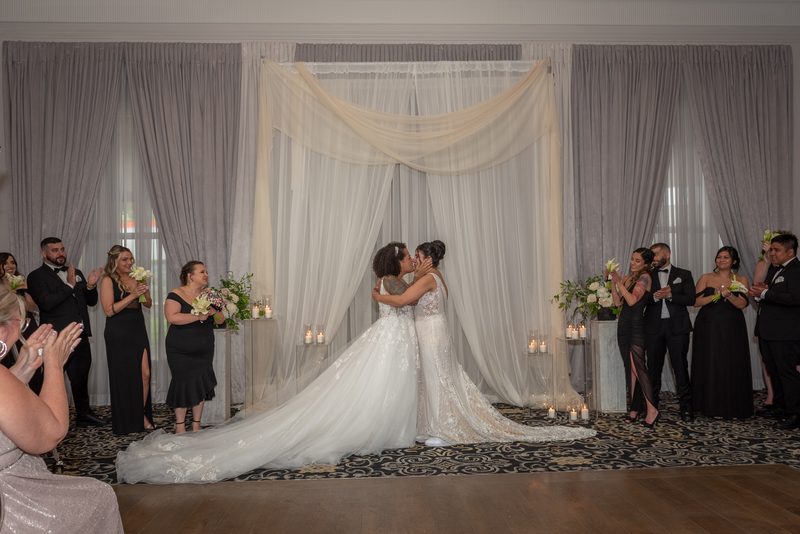 Two brides, Alexa and Nazareth, share an intimate kiss at the center of a draped archway during their indoor ceremony.