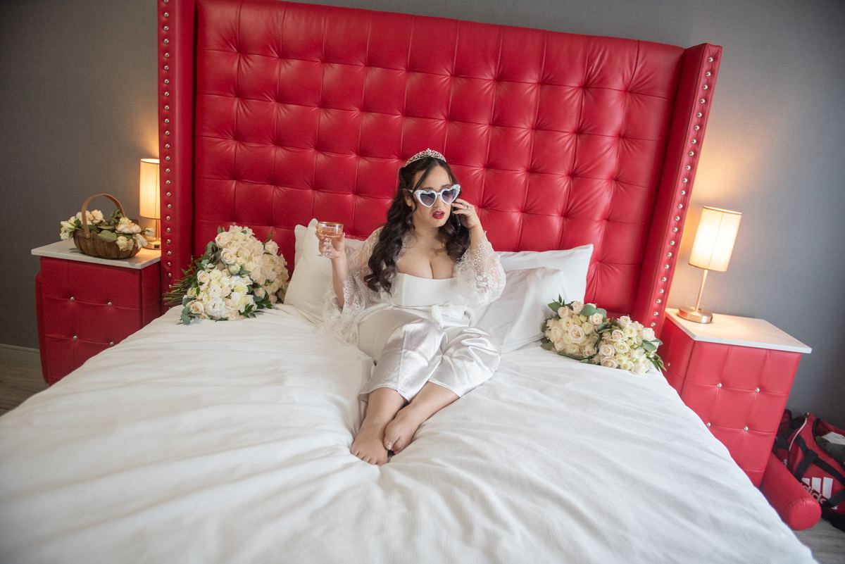 A bride relaxes on a red tufted bed with wine and heart-shaped sunglasses, surrounded by white flowers.
