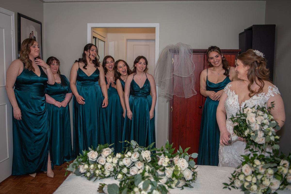 Bride in white lace gown laughs with bridesmaids in teal dresses, standing near a wooden door and veil.