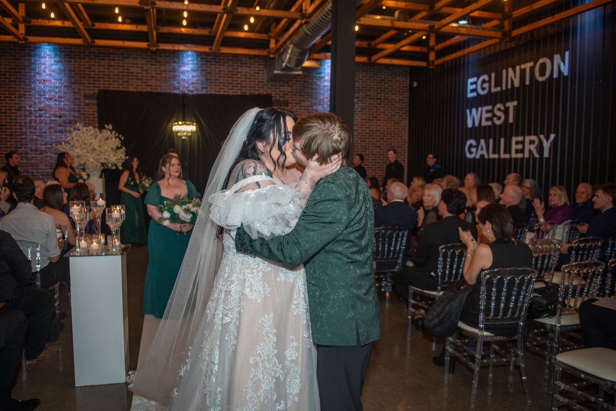 A bride and groom kiss during their wedding ceremony, surrounded by guests in clear chairs, amid exposed brick.