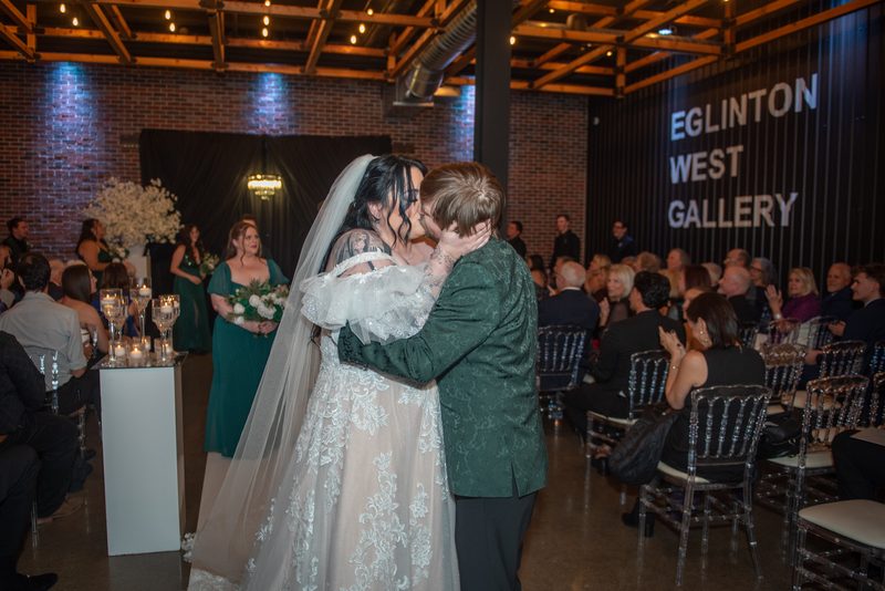 A bride and groom kiss during their wedding ceremony, surrounded by guests in clear chairs, amid exposed brick.