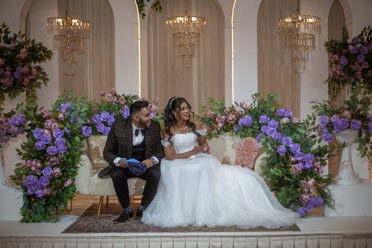A smiling newlywed couple sits together on a decorated stage under elegant chandeliers and floral arrangements.