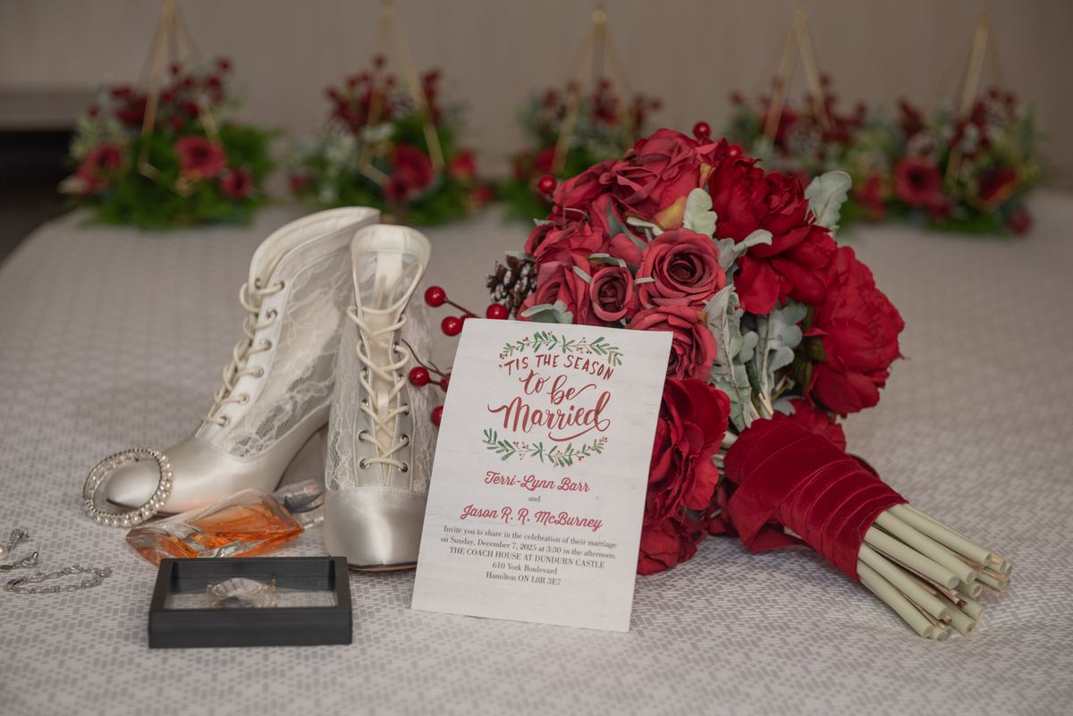 Red rose bouquet, silver lace boots, wedding invitation card, perfume bottle, and jewelry box arranged on textured surface.