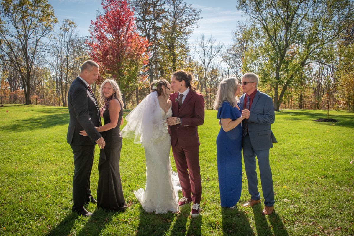 A bride and groom kiss in an autumn field surrounded by family, with golden sunlight casting long shadows.