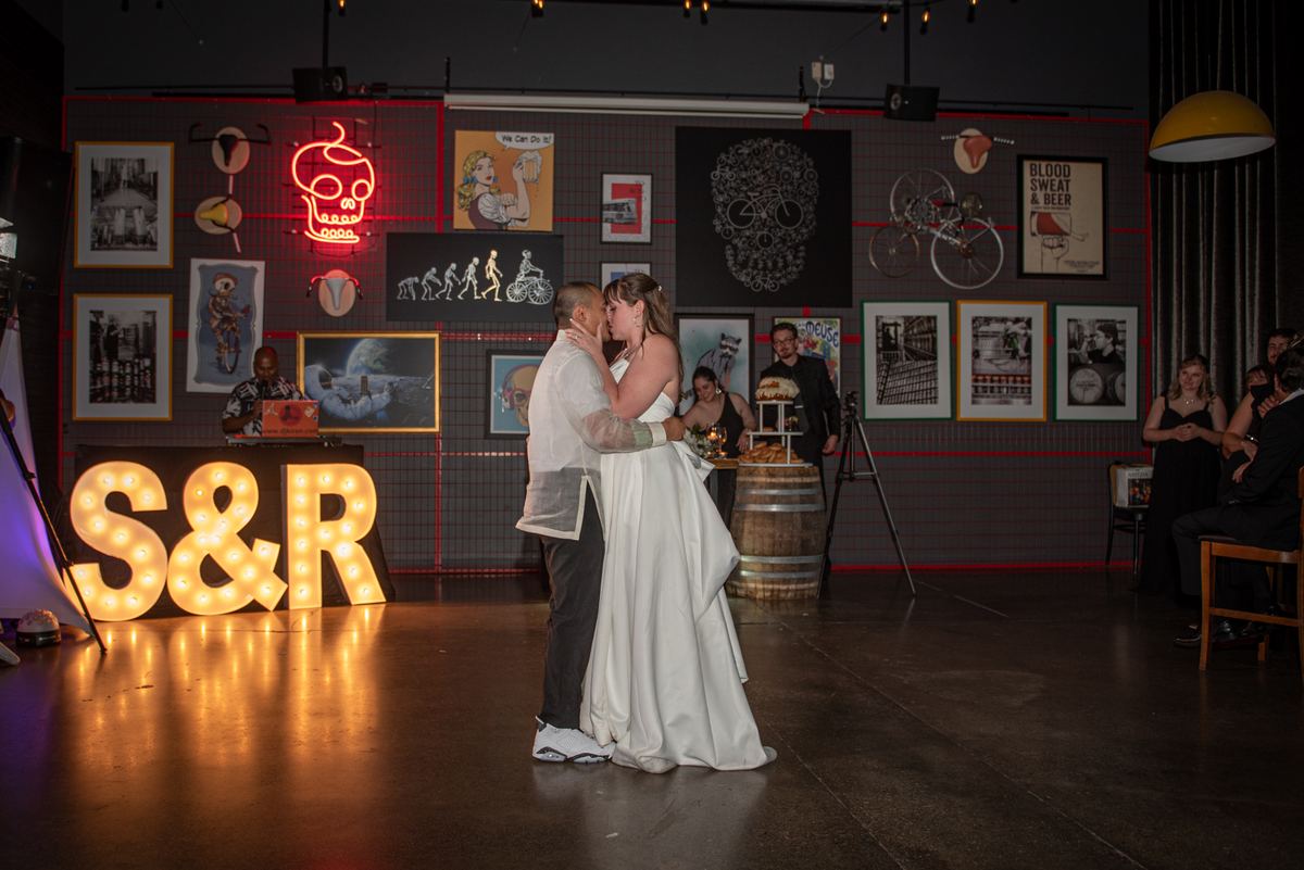 A bride and groom dance together in a warmly lit space decorated with vintage posters, a neon skull, and illuminated letters.