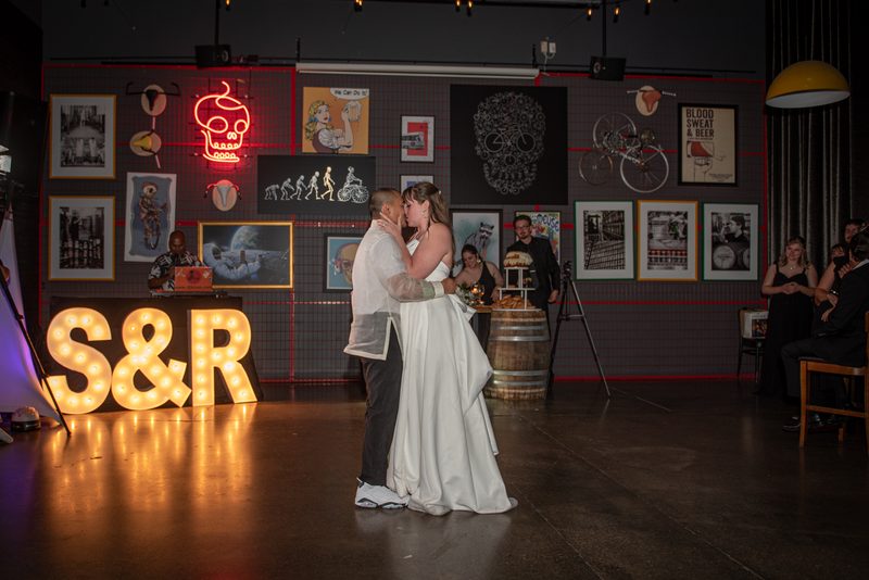 A bride and groom dance together in a warmly lit space decorated with vintage posters, a neon skull, and illuminated letters.