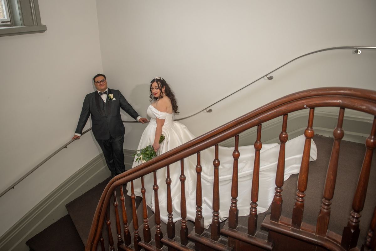 Bride and groom descend staircase together, she holding bouquet and glancing back, he leaning against railing.