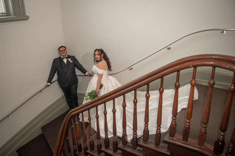 Bride and groom descend staircase together, she holding bouquet and glancing back, he leaning against railing.