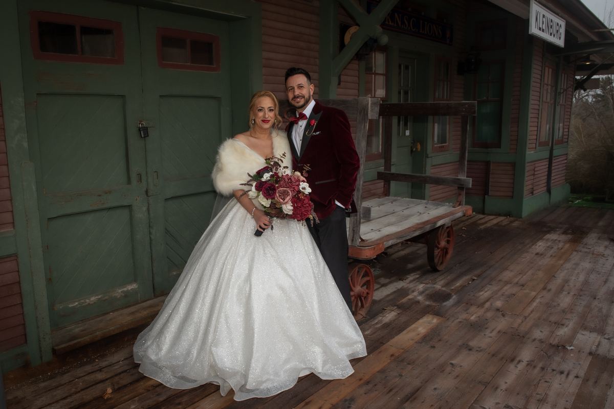 A bride in a sparkling gown and fur stole with her groom in a velvet burgundy tuxedo pose near an old train station platform.