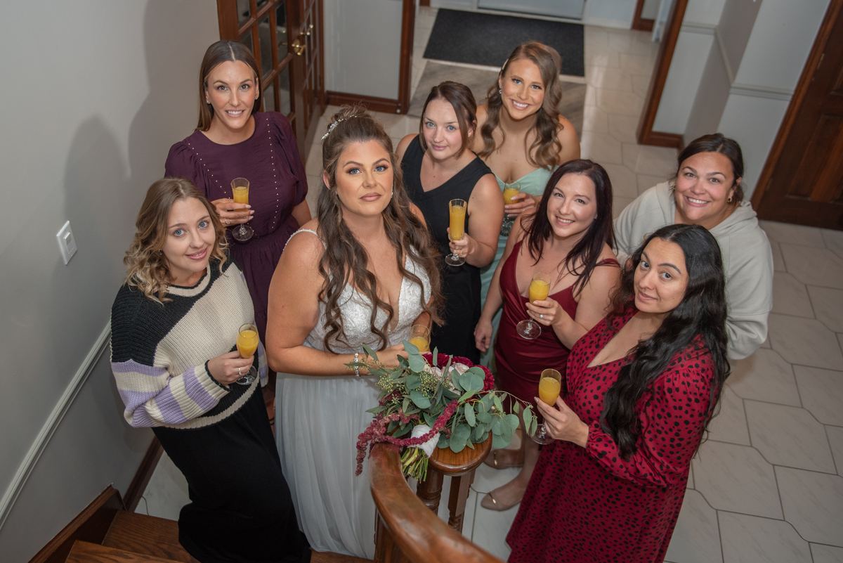 Bride and bridesmaids gather on staircase for toast, holding orange juice glasses, smiling warmly in colorful attire.