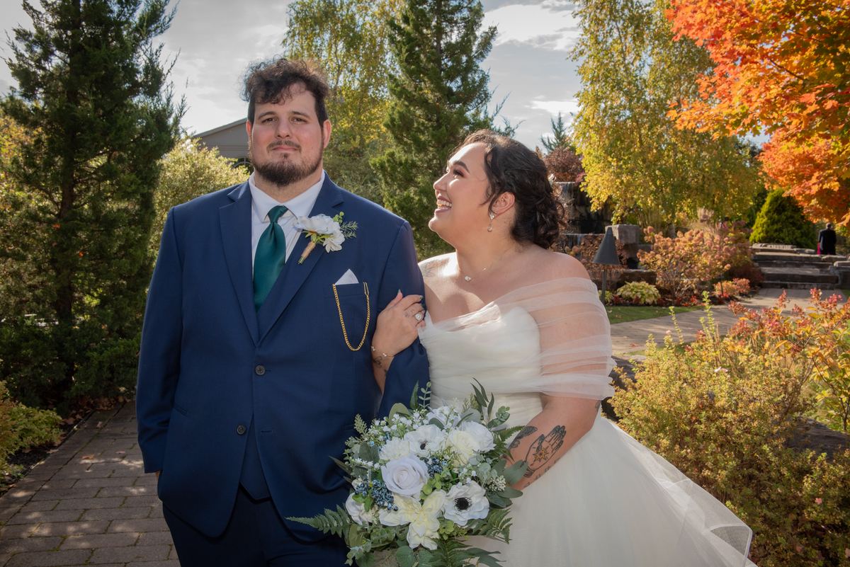 A smiling bride and her groom stand in an autumn garden, illuminated by warm sunlight filtering through colorful trees.