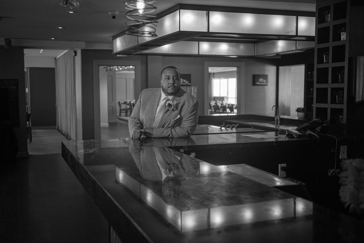 A groom in a suit stands at a sleek, reflective hotel bar, gazing directly at the camera in dramatic monochrome lighting.