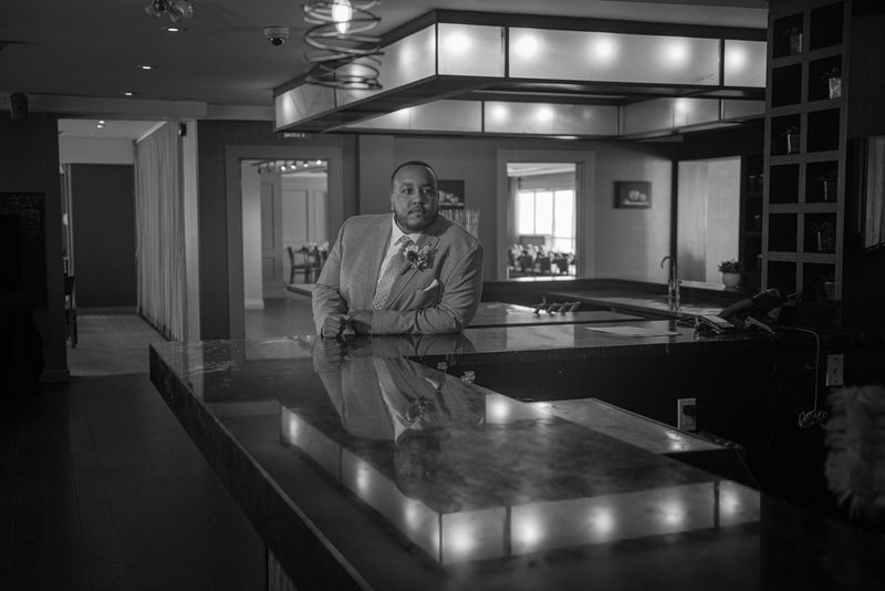 A groom in a suit stands at a sleek, reflective hotel bar, gazing directly at the camera in dramatic monochrome lighting.