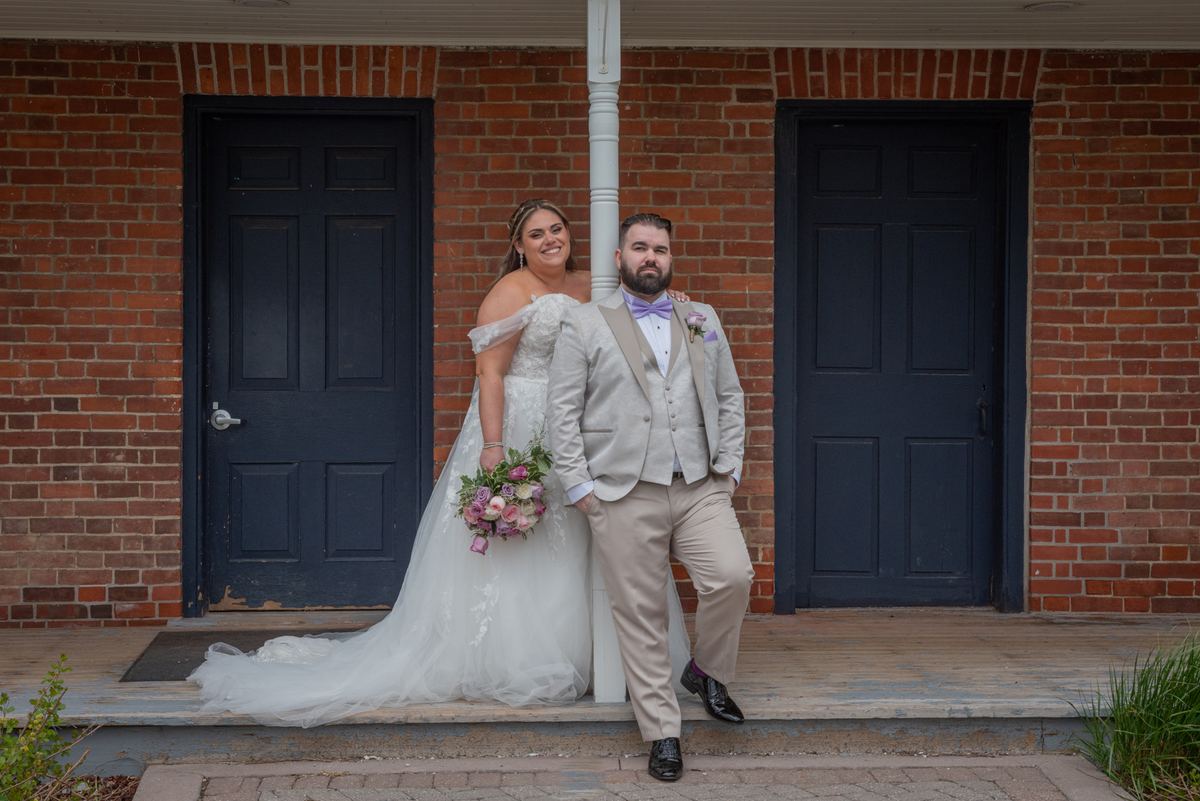 Bride in white dress and pink flowers stands with groom in dark suit on porch of red brick building, leaning against post.