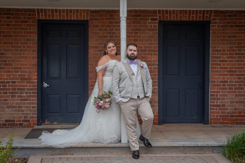 Bride in white dress and pink flowers stands with groom in dark suit on porch of red brick building, leaning against post.