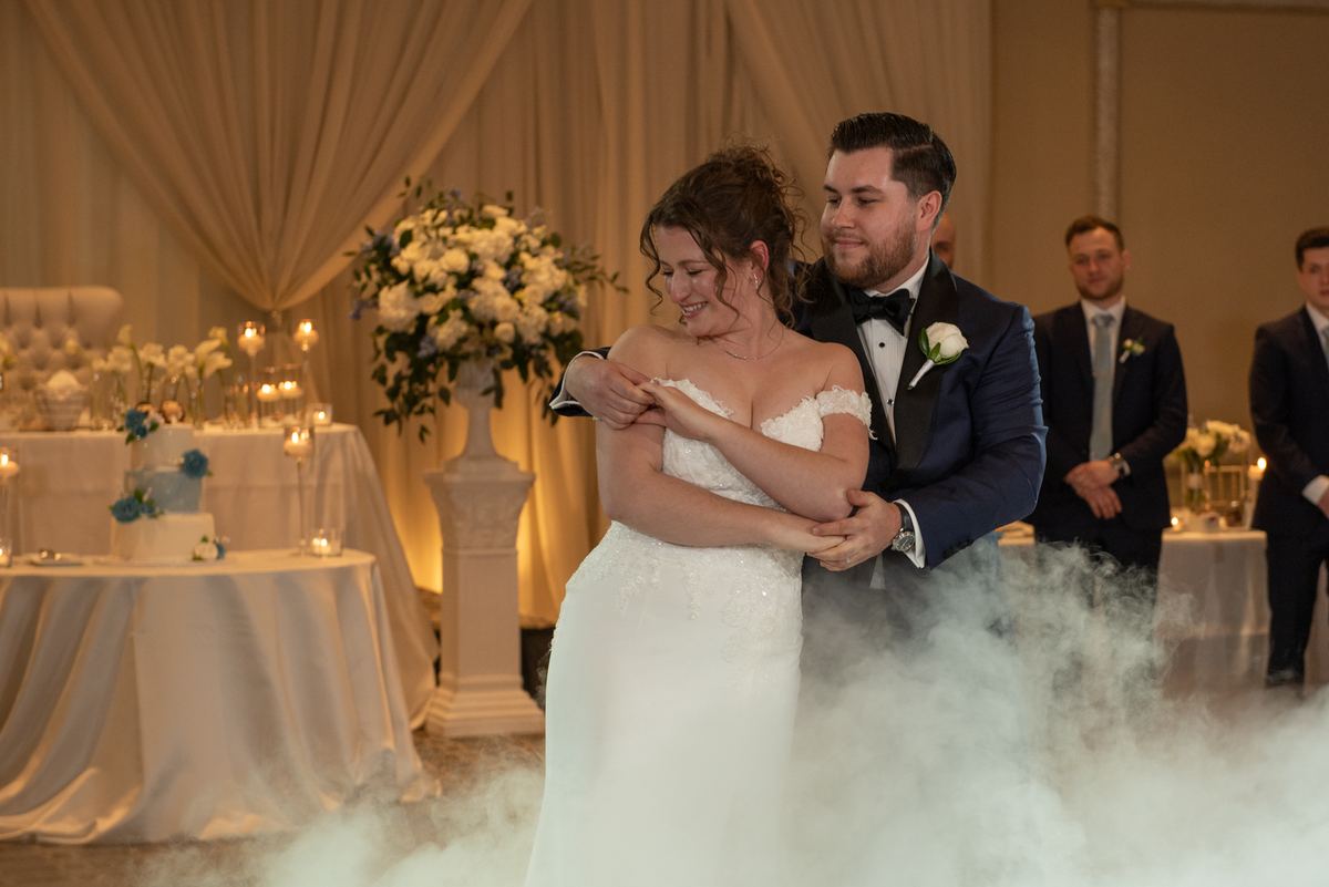 Bride and groom dance intimately, surrounded by fog and warm lighting, with groomsmen in background.