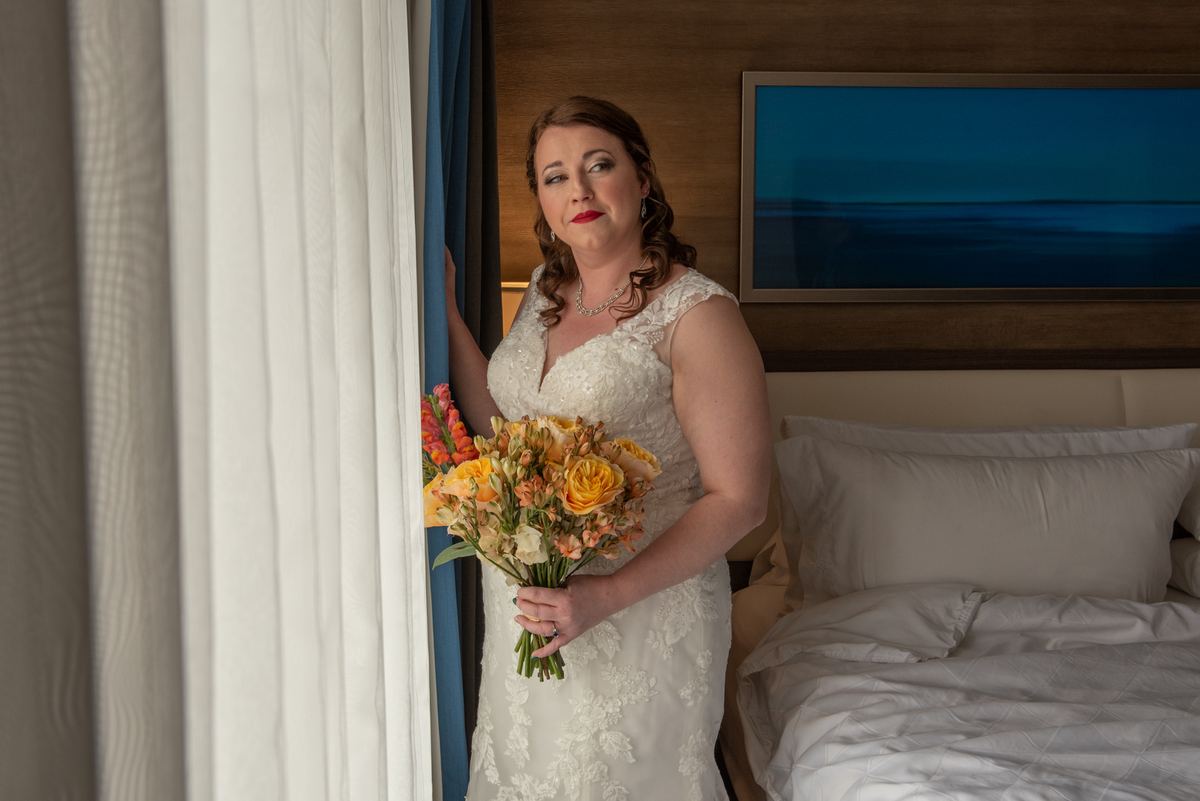 Bride in white lace gown holds orange and yellow flowers near window, light filtering through curtains.