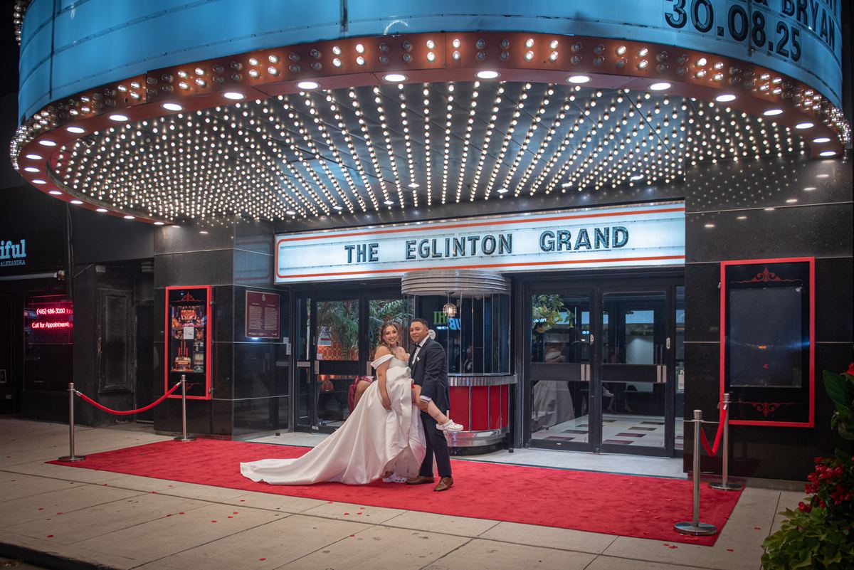 A bride and groom stand on a red carpet outside a cinema at night, illuminated by warm lights from the marquee.