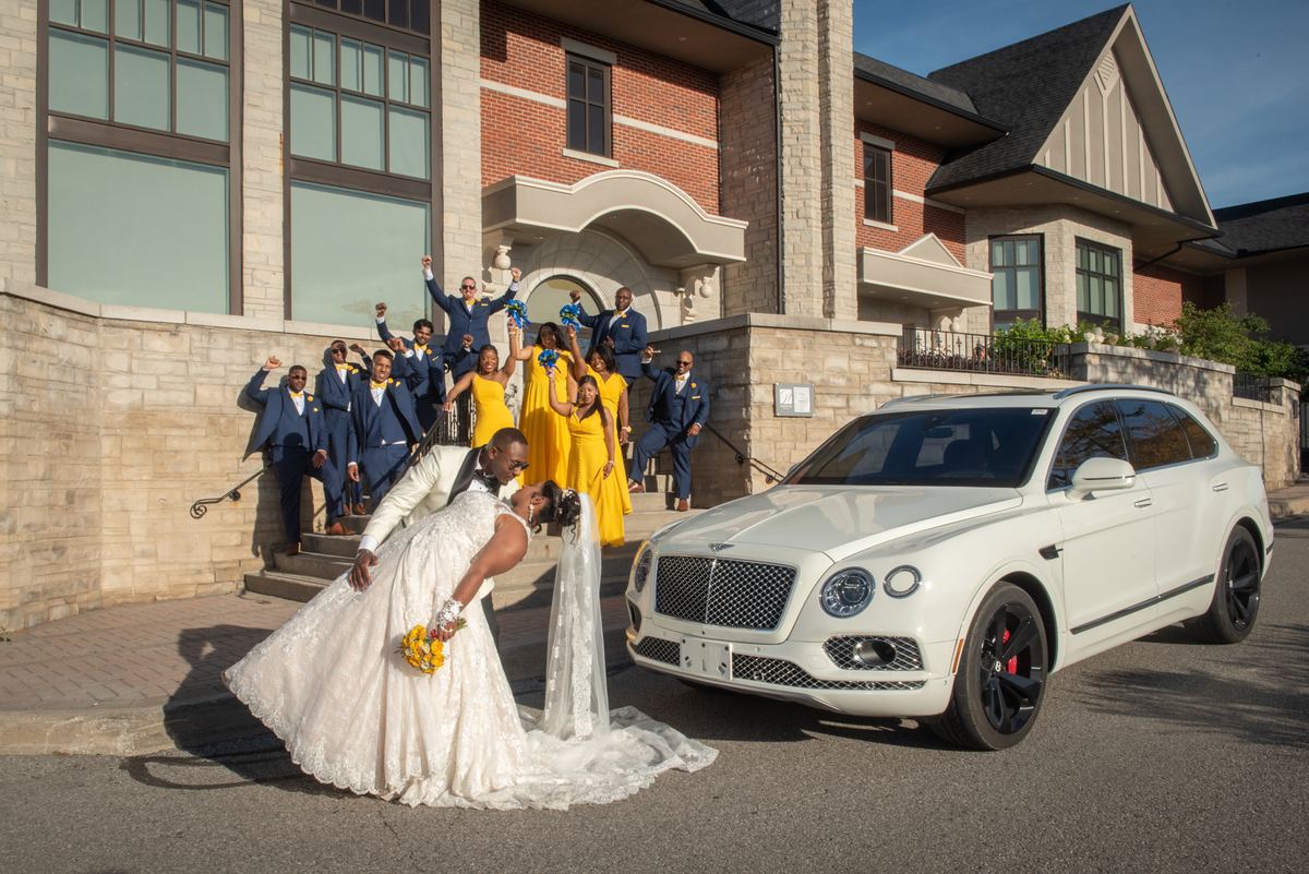Bride and groom pose playfully with wedding party on steps of stately home as sunlight highlights Bentley SUV.