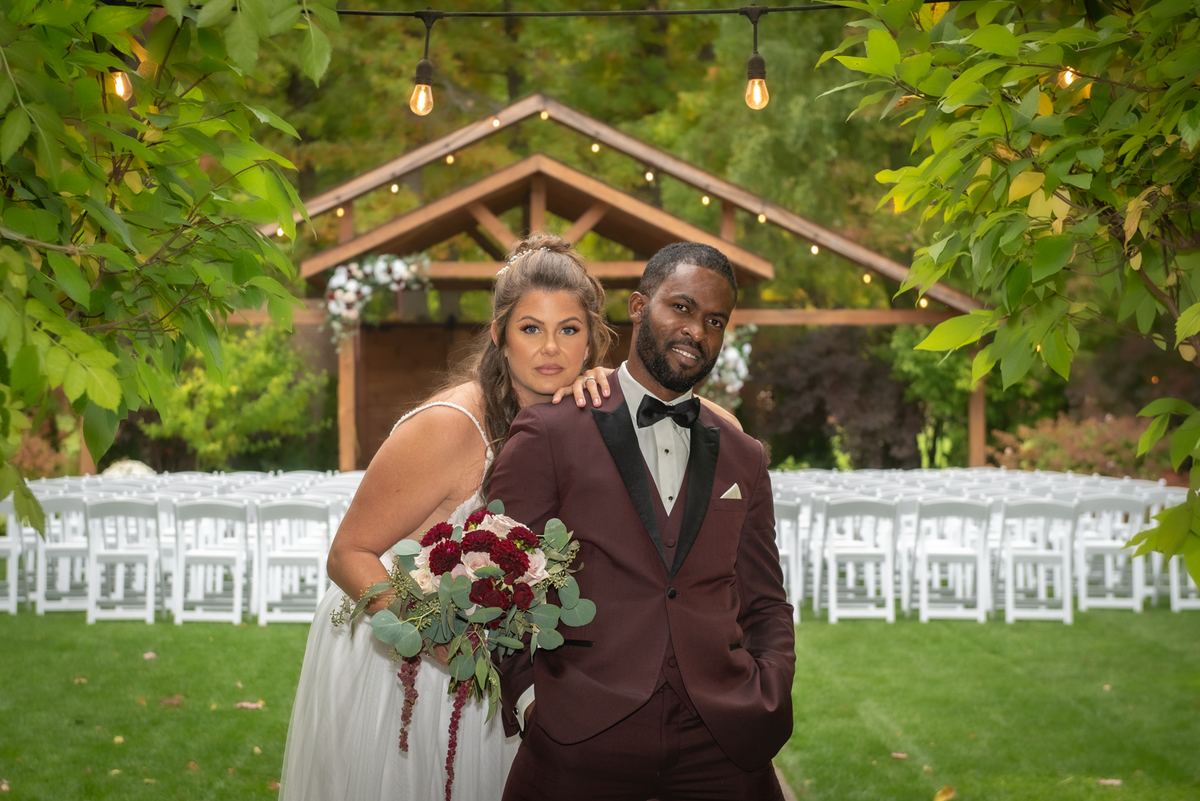 Bride and groom stand in an outdoor setting with greenery and string lights, framed by white chairs and wooden arbor.